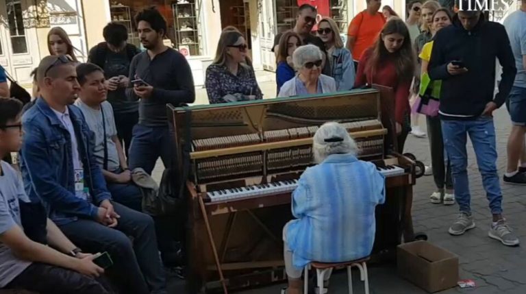 A grandma sits in front of the piano and starts playing. People passing by gathered around her to watch this wonder.